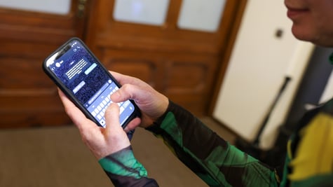 woman in green, black right top holds smartphone, shows screen with text messages, brown door