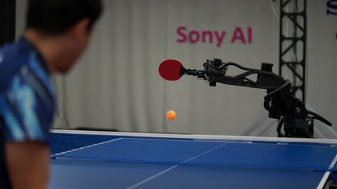 black robot hand, red racket, blue table tennis table, human player, black hair, blue shirt