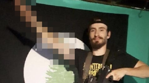 white man, black beard, moustache, salutes, black tshirt, yellow sign, brown belt, dark rifle, firn 