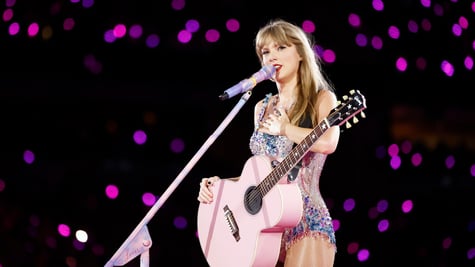 Taylor Swift, pink-violet sparkly bodysuit, pink guitar, white microphone, dark stadium crowd