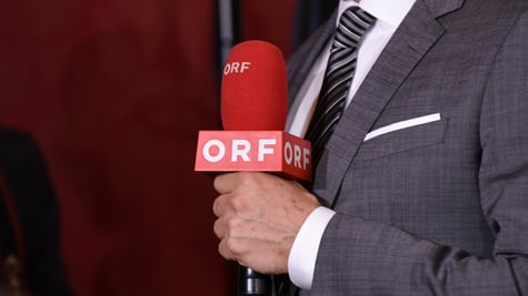 grey suit, white shirt, grey tie with stripes, dark orange microphone, ORF white letters, white hand
