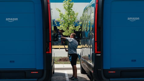 two blue amazon vans, male fat worker in between them, cleans the left one, sunny day, tree