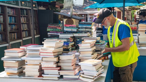 old man, live yellow reflective vest, blue tshirt, piles of books, shelves of books, tan, brown shorts