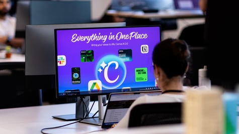 Employee at white desk, black screen frame, violet canva logo on screen, office, dark short hair