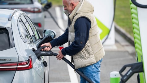 silver electric car, black charging cable, smiling old man in jeans, black jumper, sand color vest