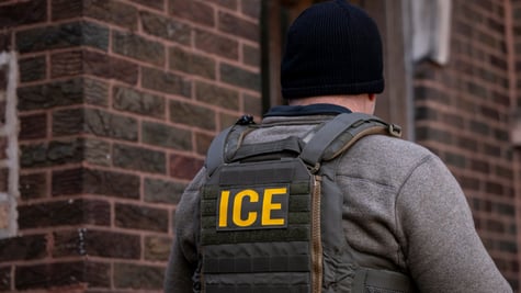 ice agent from back, black hat, white male, grey top, yellow letters ICE, dark brown brick wall 