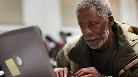 Black man in grey hair, moustache, and beard, grey laptop, army green winter jacket, 
