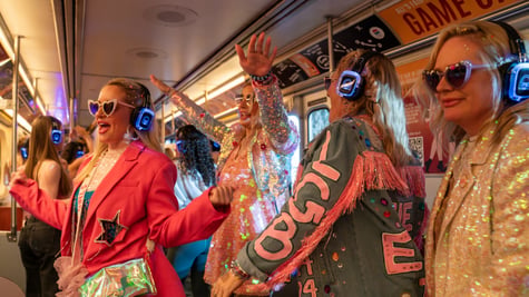 four blonde women wear blue whiny headphones, sparkly outfits, on a white bus, pink jacket