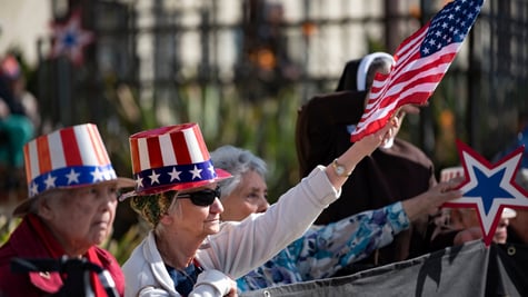 elderly white American couple, tall hat with US flag, black sunglasses, black fence, white jacket