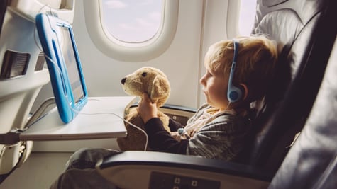 blonde boy, on flight, white plane salon, light brown teddy bear, baby blue tablet with wire