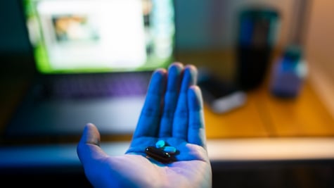 two white, two brown pills in white male hand, open laptop screen, brown desk.
