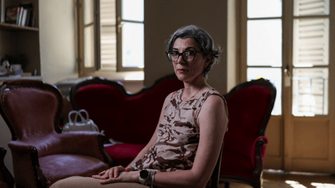 italian woman, sleevless shirt, grey hair, black frame glasses, tall windows, vintage sofa