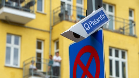 white surveillance camera on grey poll, blue sticker with palantir written on it, yellow building