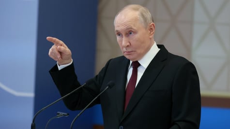 putin points right hand finger, black suit, white collar shirt, bordeaux tie, blue wall