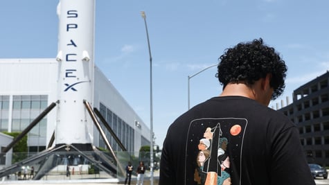spaceX white rocket, white building, black man, black t-shirt with rocket, black curly hair, blue sky 