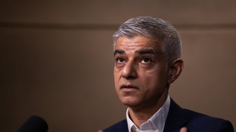 Sadiq Khan looking up, grey hair, black suit, white collar shirt, dark brown background, grey hair