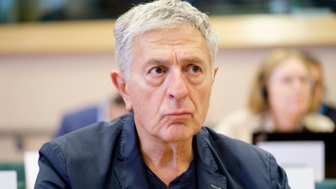 Stelios Kouloglou, white elder greek man, black collar shirt, no tie, grey hair, dark blazer,