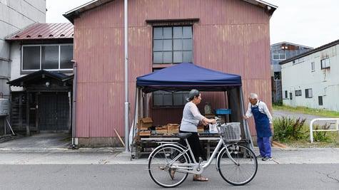 A woman pushes a bicycle past a vendor at a morning market in Gojome