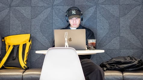 Man wearing headphones and baseball cap working on laptop at cafe table with backpack and iced coffee