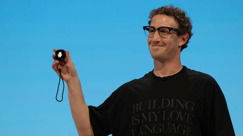 Zuckerberg, curly hair, black tshirt, baby blue background, small round gadget in raised right hand