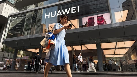 A pedestrian looks at a smartphone while walking past a Huawei retail store displaying an illuminated Huawei sign and smartphone advertisements 