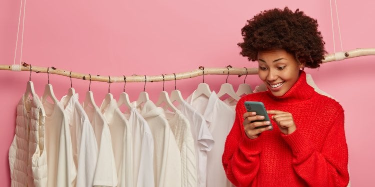 Young woman looking excitedly at cellphone and a line of white clothes hooked on a rack in the backg