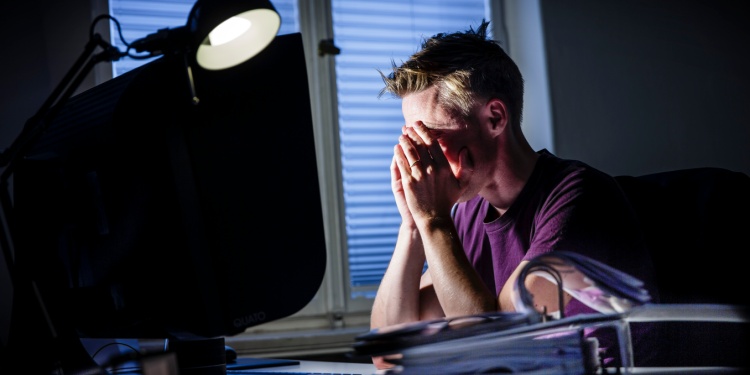 A stressed out office worker clutching his head at night, next to a bright lamp.