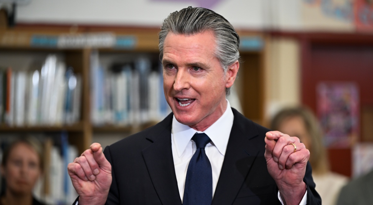 Gavin Newsom, black suit and tie, fists up, grey combed back hair, library, books, smiling, talking