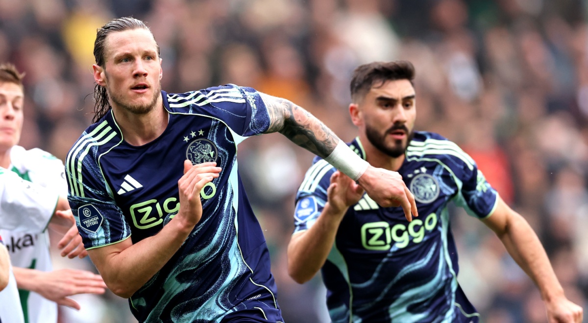 two football players run, dark blue tops, ajax logo, crowded stadium in the back, long blond hair