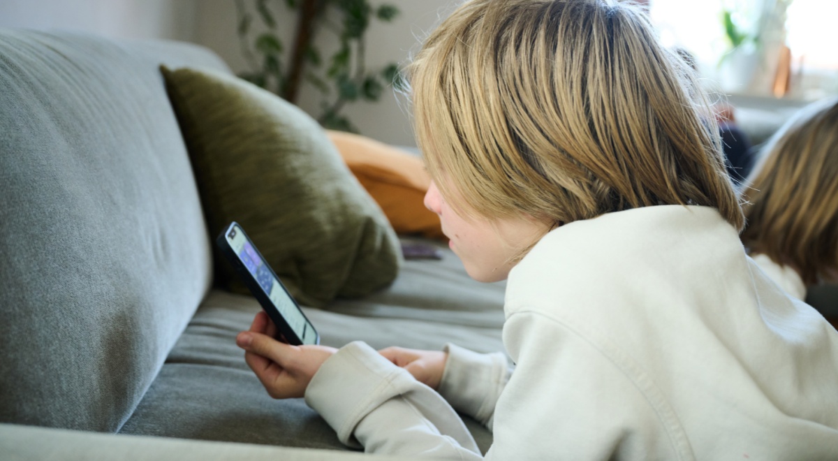 blonde boy, shoulder length hair, scrolls smartphone, white sweatshirt, grey sofa