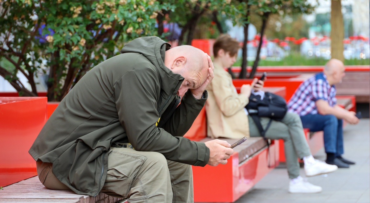 bald man, scrolls phone, red bench, dark green jacket, green trees