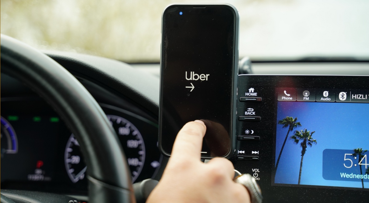 uber app open on black smartphone screen, car screen, blue sky and palm tree, driving wheel