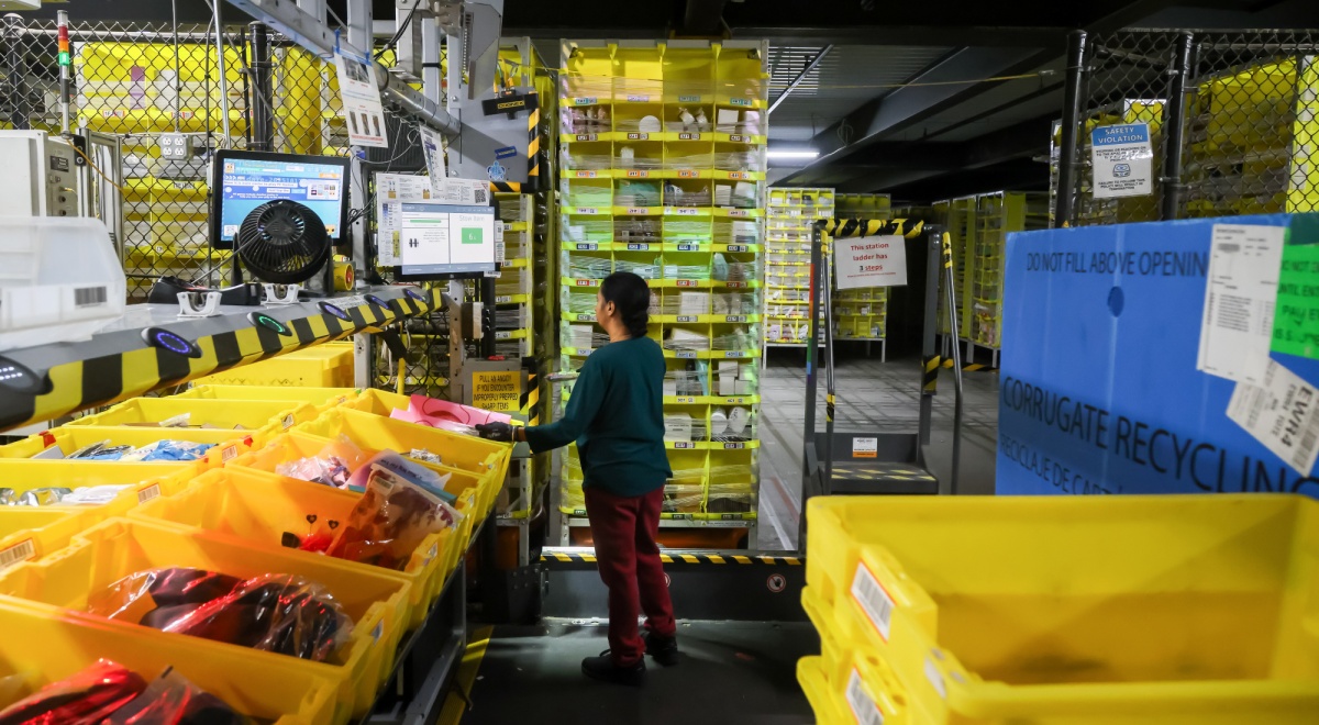 Female worker, dark hair, Amazon fulfillment center, yellow parcel boxes, fence, dark red throusers