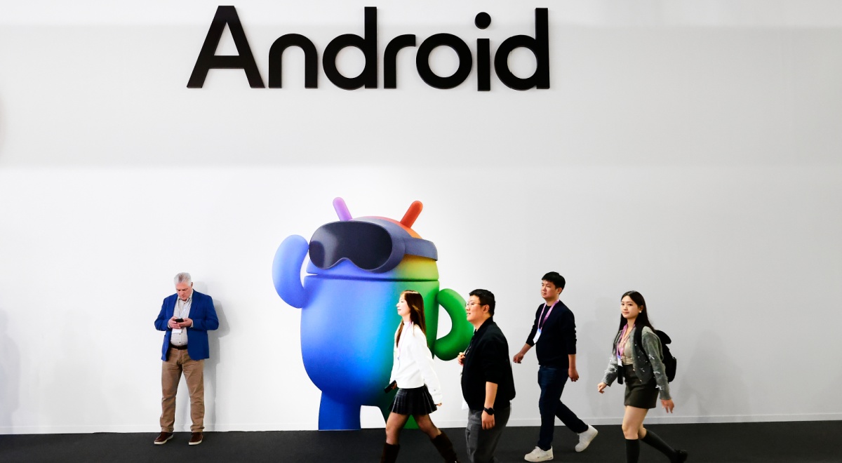 three asians walk pass blue android mascot, white wall, one man leaning to wall, Android black sign