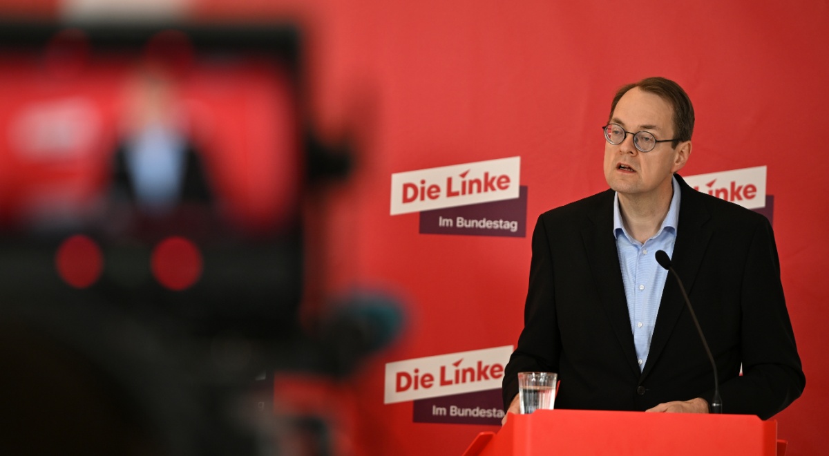Soeren Pellmann, man in black suit, light blue shirt, no tie, collar unbuttoned, red photo wall