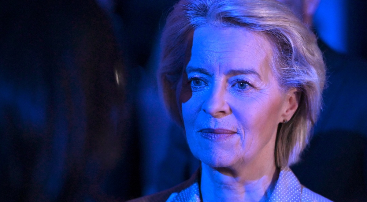 Ursula von der Leyen, blonde woman, semi-smile, white skin lit up in dark blue light
