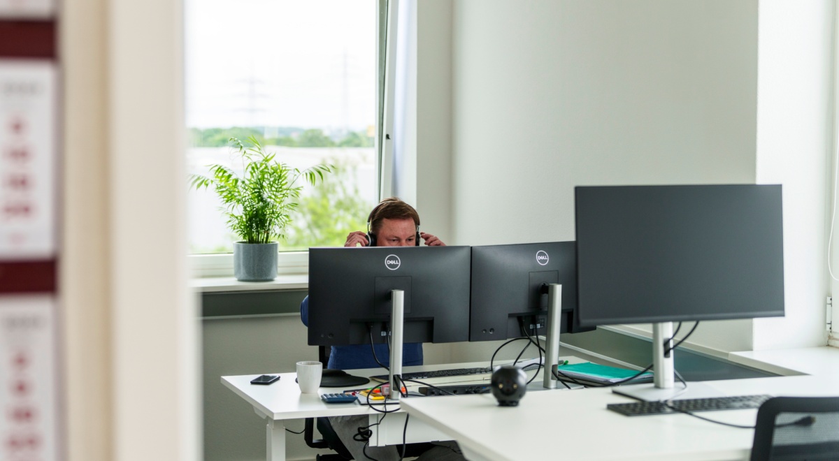 office worker with headset, hands up, crear window, green tree, three dell monitors, white desks 