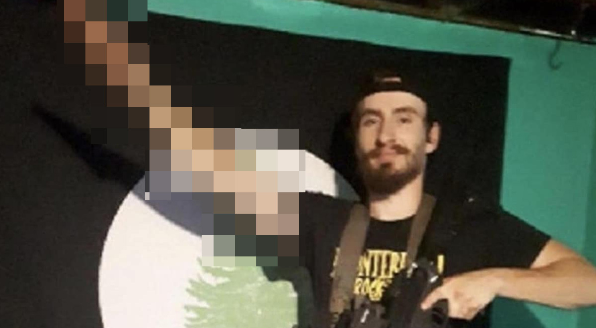 white man, black beard, moustache, salutes, black tshirt, yellow sign, brown belt, dark rifle, firn