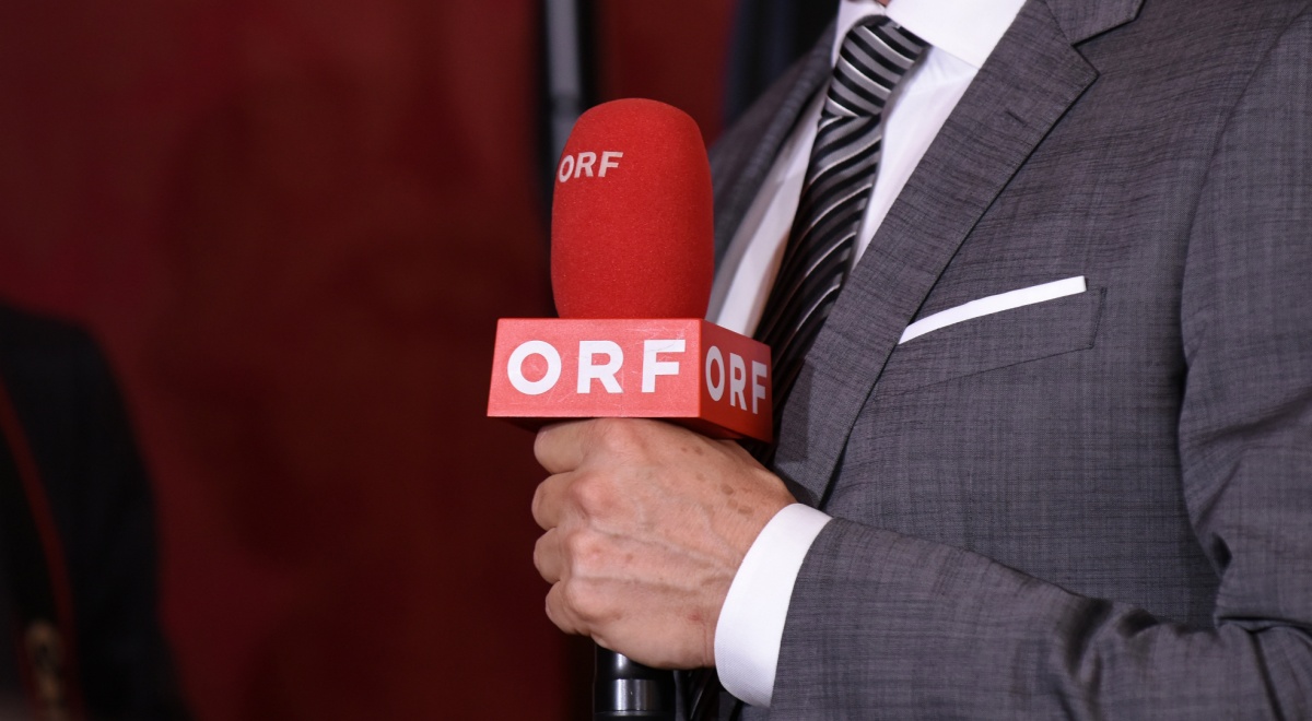 grey suit, white shirt, grey tie with stripes, dark orange microphone, ORF white letters, white hand