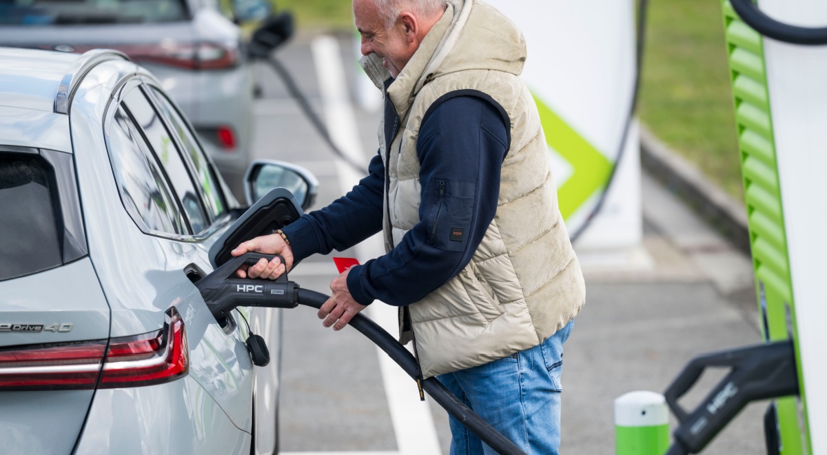 silver electric car, black charging cable, smiling old man in jeans, black jumper, sand color vest