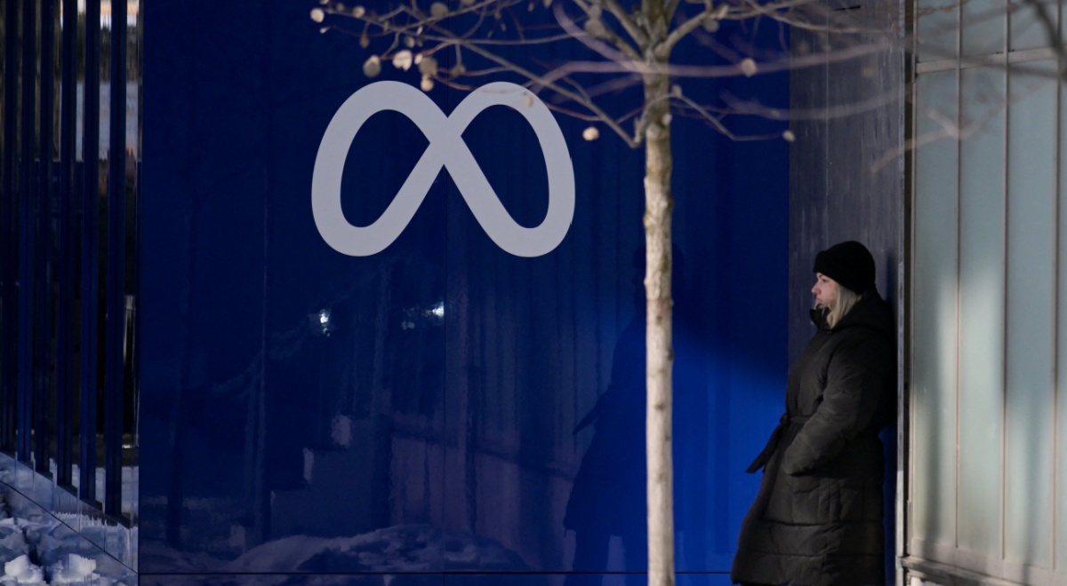 dark grey meta logo on dark blue wall, winter, woman in long black winter coat, blonde,