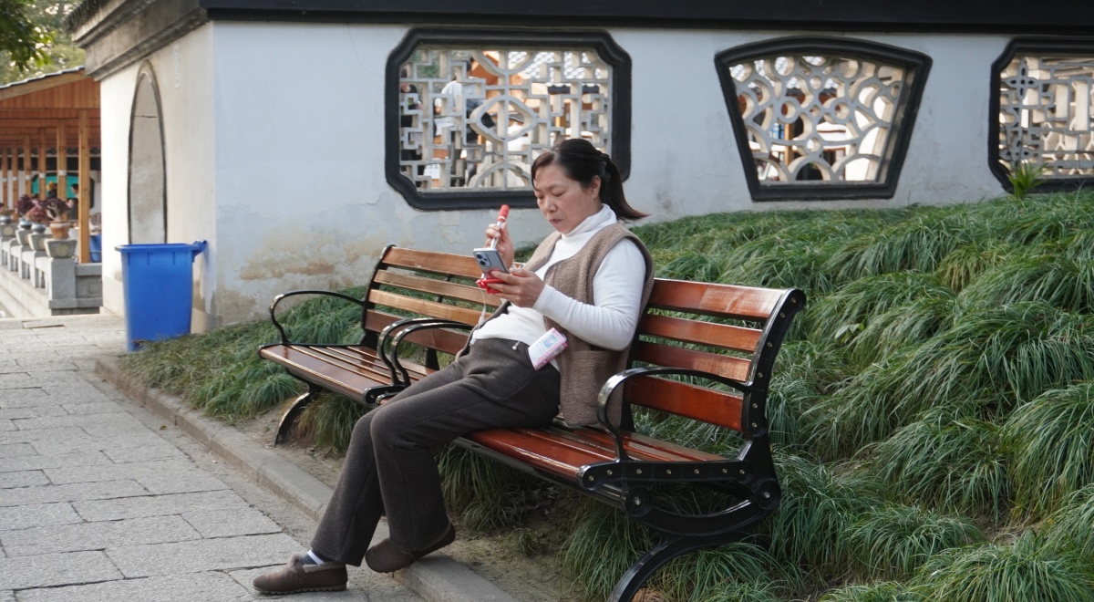 old chinese woman, black hair, wooden bench, smartphone, white building wall, green bushes