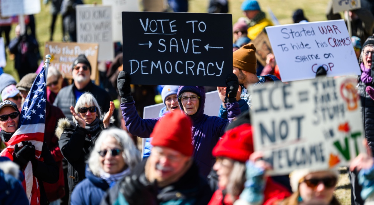 crowd of people holding protest signs, black sign, white letters, vote to save democracy