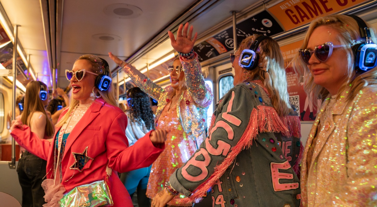 four blonde women wear blue whiny headphones, sparkly outfits, on a white bus, pink jacket