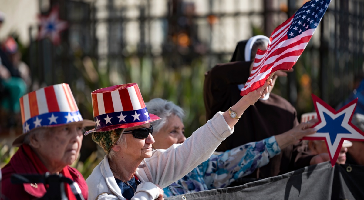 elderly white American couple, tall hat with US flag, black sunglasses, black fence, white jacket