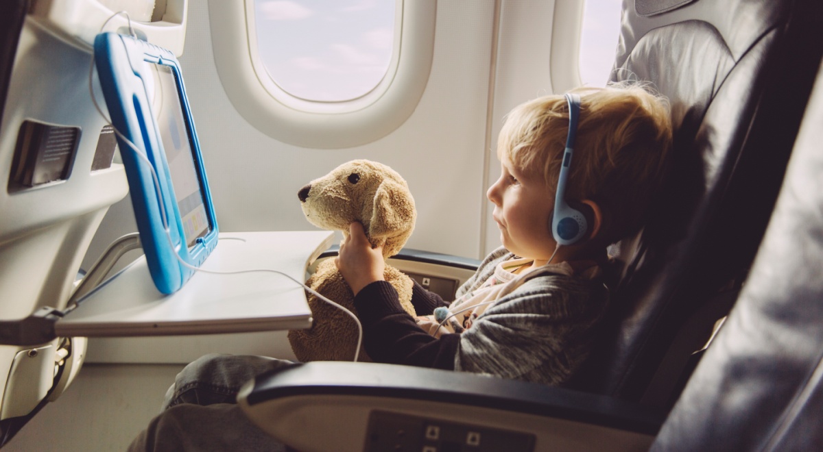 blonde boy, on flight, white plane salon, light brown teddy bear, baby blue tablet with wire