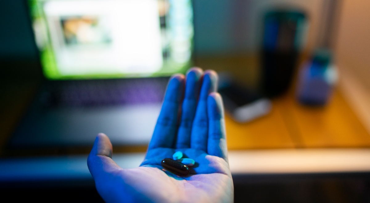 two white, two brown pills in white male hand, open laptop screen, brown desk.