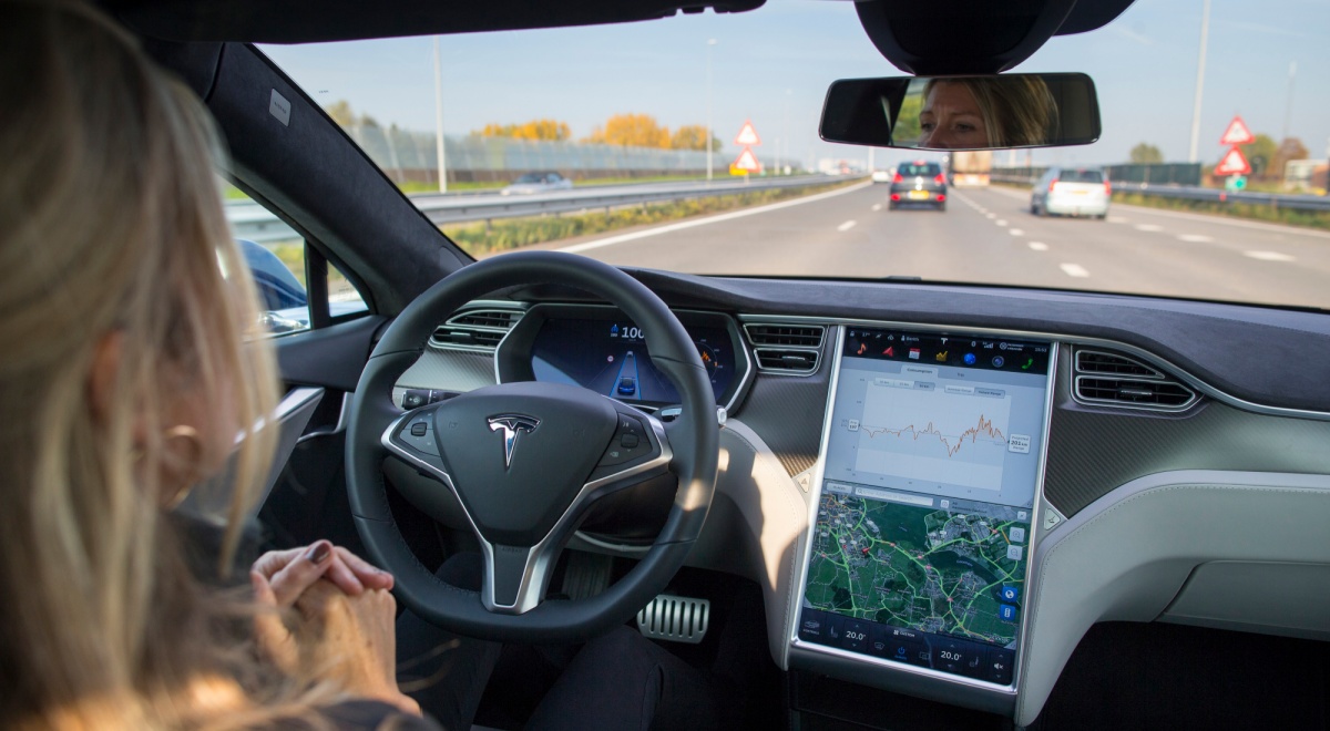 blue sky, inside of car, road ahead ,female blonde driver, tesla car, T on driving wheel, big screen