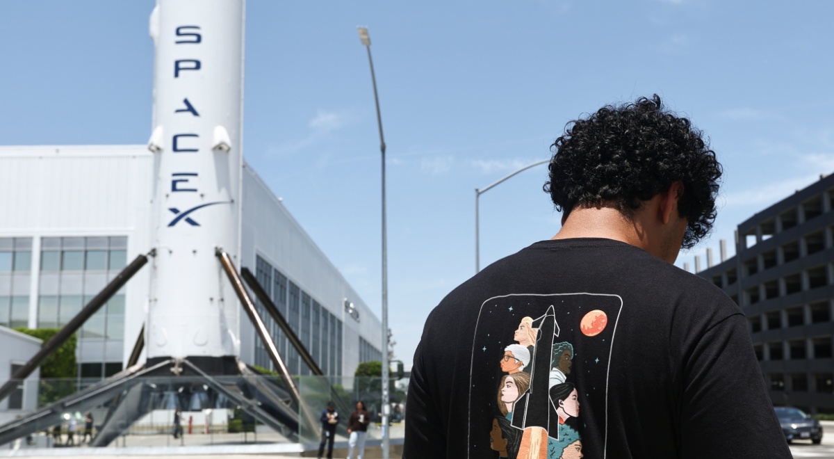 spaceX white rocket, white building, black man, black t-shirt with rocket, black curly hair, blue sky 