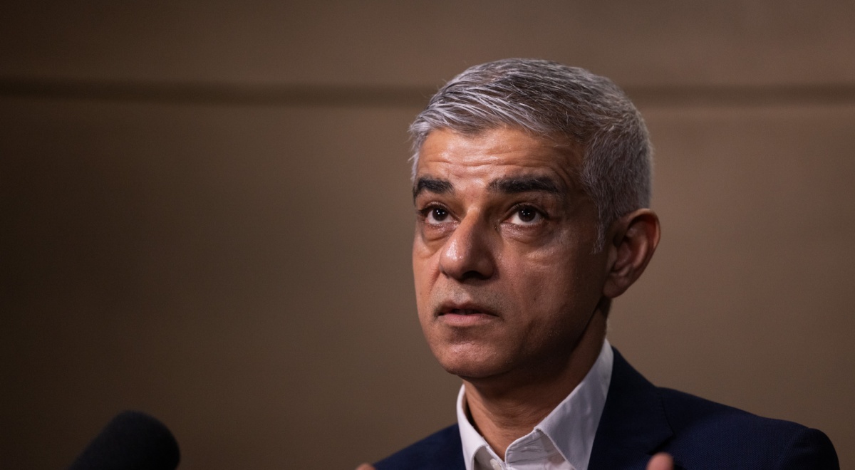 Sadiq Khan looking up, grey hair, black suit, white collar shirt, dark brown background, grey hair